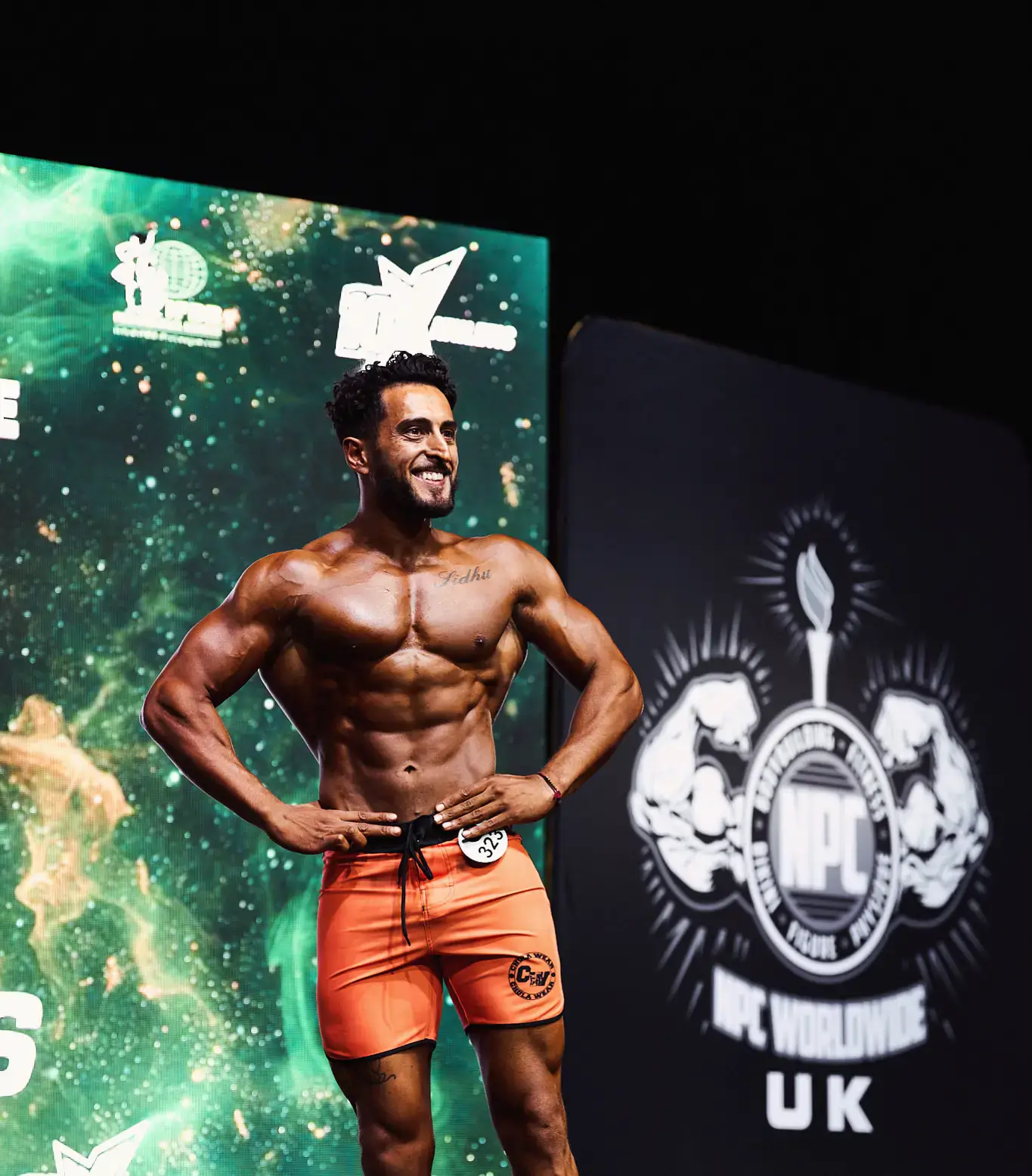 Sidhu Fitness founder Tony Sidhu competing on a bodybuilding stage in orange shorts